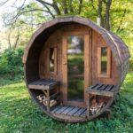 Eine rustikale Fasssauna aus Holz steht inmitten einer grünen Naturkulisse mit Bäumen und Gras.